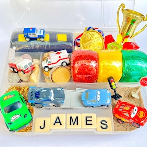 Pu&ograve; includere: Un contenitore di plastica trasparente pieno di macchinine giocattolo, un trofeo, una medaglia e plastilina colorata. Il nome "JAMES" &egrave; scritto con tessere. Le auto sono di vari colori e stili, tra cui un'autopompa e un'ambulanza.
