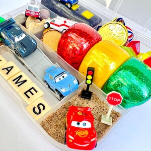 Pu&ograve; includere: Un contenitore di plastica trasparente pieno di macchinine giocattolo, plastilina e altri piccoli giocattoli. Il contenitore include un'auto da corsa rossa, un'auto blu, un segnale di stop e un semaforo. Il nome "JAMES" &egrave; scritto con lettere.