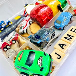 Pu&ograve; includere: Un organizer di plastica trasparente pieno di macchinine giocattolo, un trofeo d'oro, una medaglia d'oro e frutta decorativa. Le auto sono di vari colori, tra cui verde, blu e nero. Il nome "JAMES" &egrave; scritto con lettere.
