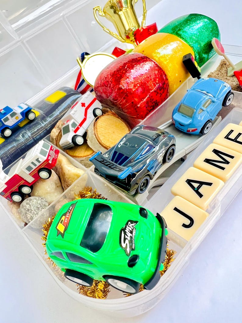 Pu&ograve; includere: Un organizer di plastica trasparente pieno di macchinine giocattolo, un trofeo d'oro, una medaglia d'oro e frutta decorativa. Le auto sono di vari colori, tra cui verde, blu e nero. Il nome "JAMES" &egrave; scritto con lettere.
