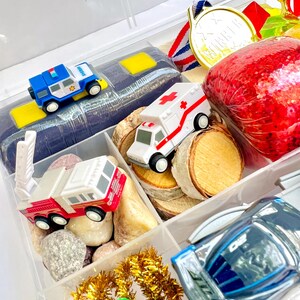 Pu&ograve; includere: Un contenitore di plastica trasparente diviso contenente veicoli giocattolo, tra cui un'auto della polizia blu e bianca, un'ambulanza bianca con una croce rossa e un camion dei pompieri rosso. Include anche una medaglia d'oro, fette di legno e un oggetto rosso scintillante.