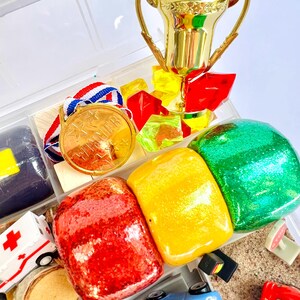 Pu&ograve; includere: Un trofeo d'oro, una medaglia d'oro con la scritta "WINNER" e oggetti colorati, glitterati e giocattolo sono disposti in un contenitore di plastica trasparente. Gli oggetti includono pietre glitterate rosse, gialle e verdi, un'ambulanza giocattolo e macchinine.