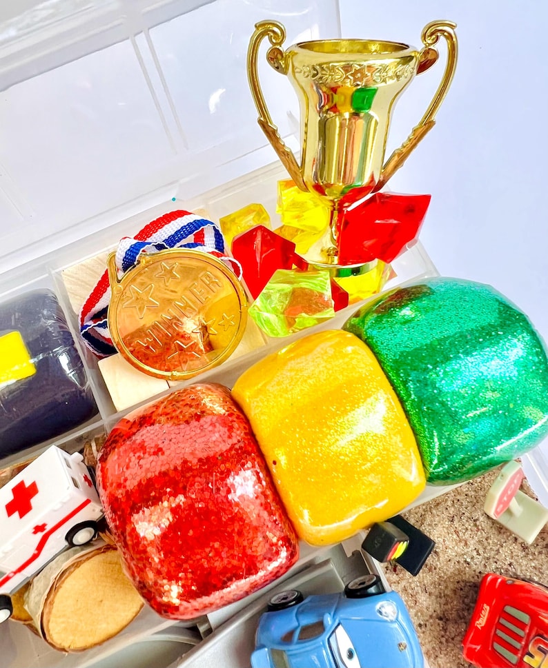 Pu&ograve; includere: Un trofeo d'oro, una medaglia d'oro con la scritta "WINNER" e oggetti colorati, glitterati e giocattolo sono disposti in un contenitore di plastica trasparente. Gli oggetti includono pietre glitterate rosse, gialle e verdi, un'ambulanza giocattolo e macchinine.