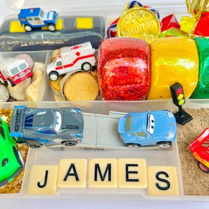 Pu&ograve; includere: Un contenitore di plastica trasparente pieno di macchinine, un'auto della polizia, un'ambulanza e un camion dei pompieri. Il nome "JAMES" &egrave; scritto con tessere. Altri oggetti includono una medaglia d'oro, oggetti rossi e gialli glitterati e un semaforo.