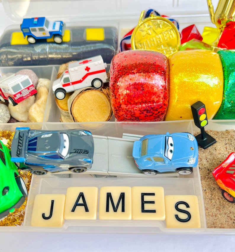 Pu&ograve; includere: Un contenitore di plastica trasparente pieno di macchinine, un'auto della polizia, un'ambulanza e un camion dei pompieri. Il nome "JAMES" &egrave; scritto con tessere. Altri oggetti includono una medaglia d'oro, oggetti rossi e gialli glitterati e un semaforo.