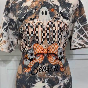 May include: A short-sleeve t-shirt with a black and tan bleached tie-dye pattern. The graphic features a white ghost, black bats, and the text SPOOKY SEASON in a checkered font, accented by an orange and white checkered bow and white flowers.