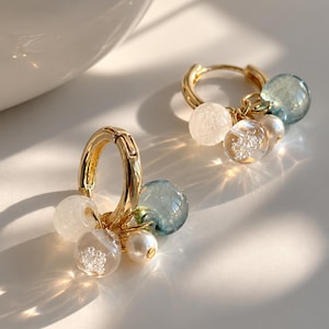 May include: A pair of gold hoop earrings, each featuring a cluster of dangling beads. The charms include a translucent white bead, a clear bead with internal white flecks, a small white pearl-like bead, and a larger translucent blue bead.