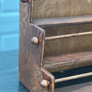 May include: A rustic, wooden spice rack with three shelves and a back panel. The rack is stained a dark brown color, with visible wood grain. Small, round knobs are attached to the sides. The shelves are supported by wooden dowels.