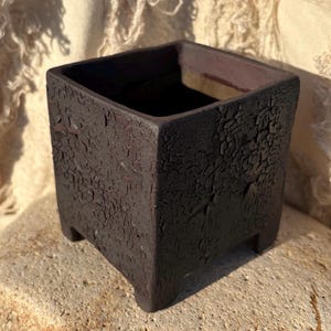 Puede incluir: Una maceta cuadrada de cerámica marrón oscuro con una superficie texturizada y agrietada. La maceta tiene un interior profundo y se asienta sobre cuatro pequeños pies. La estética general es rústica y terrosa.