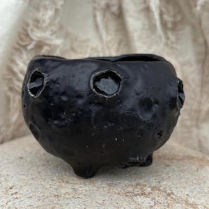 Może przedstawiać: Mała, czarna ceramiczna miska o fakturze i trzech małych nóżkach. Misa ma trzy okrągłe otwory z odblaskowym, ciemnym wnętrzem. Misa stoi na jasnej, teksturowanej powierzchni.