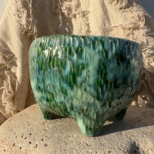 May include: A handcrafted, ceramic planter with a textured surface and three small feet. The planter features a glossy glaze in shades of green and teal. The round, open-top container is suitable for small plants.