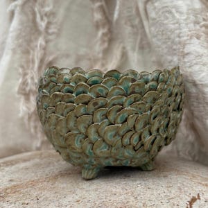 May include: A handcrafted ceramic bowl with a textured, scale-like pattern in shades of green and brown. The bowl is supported by three small feet and has a scalloped rim. The background is a soft, neutral fabric.