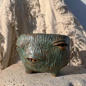 Può includere: Un vaso di ceramica verde acqua e marrone con un design a forma di volto. Il vaso ha tre occhi e una superficie strutturata, appoggiato su una pietra. Il vaso ha una forma quadrata ed è sostenuto da piccoli piedini. Lo sfondo presenta un tessuto strutturato bianco sporco.