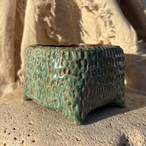 May include: A set of three ceramic mugs with a speckled, matte finish. The mugs are in shades of cream, light brown, and dark brown. Each mug has a simple, minimalist design and a rounded handle.