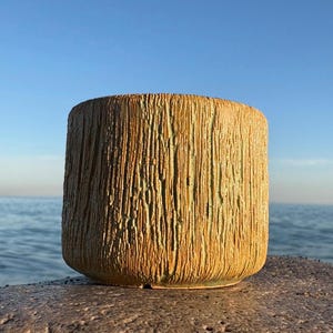 Może przedstawiać: Cylindryczny ceramiczny garnek z teksturowaną, pionowo wyłożoną powierzchnią. Garnek ma ciepły, ziemisty kolor z nutami zieleni i stoi na kamiennej powierzchni. Tło przedstawia czyste błękitne niebo i ocean.