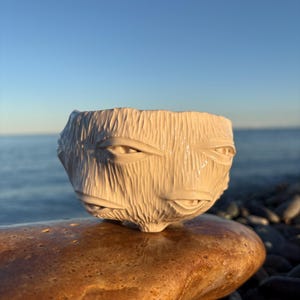 May include: A white ceramic bowl with a carved face design, resting on a brown stone. The bowl has a textured surface and features eyes, a nose, and a mouth. The background shows a blue sky and water.