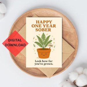 Könnte beinhalten: Eine Grußkarte mit dem Text "HAPPY ONE YEAR SOBER" und einer Cartoon-Pflanze in einem Topf. Die Karte liegt auf einem Holztablett mit einem Umschlag. Auf der Karte steht auch "Look how far you've grown."