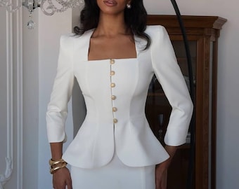 Pode incluir: Um fato de saia branco com decote quadrado e botões dourados. O casaco tem um design peplum e a saia tem uma fenda. As mangas são de três quartos. Pulseiras douradas adornam o pulso.