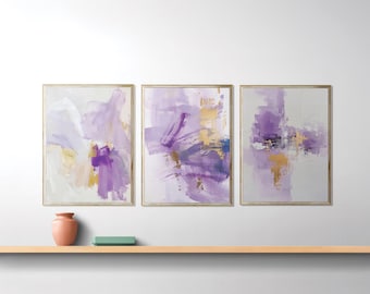 Puede incluir: Tres impresiones artísticas abstractas enmarcadas con marcos dorados, con pinceladas de color púrpura, blanco y dorado sobre un fondo gris claro. Un jarrón color melocotón y un objeto rectangular verde se encuentran en una estantería de madera.