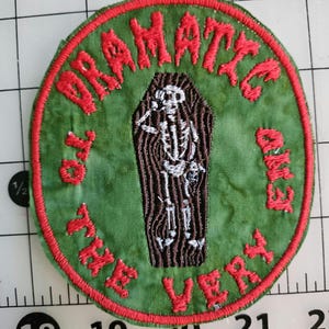May include: A round, green patch with red embroidered text and border. The text reads "DRAMATIC END TO THE VERY". A white skeleton is inside a brown coffin in the center of the patch. The patch is on a white and black grid.