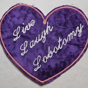 May include: A heart-shaped patch with the words "Live Laugh Lobotomy" embroidered in white script on a textured purple fabric. The patch is outlined with a pink border.