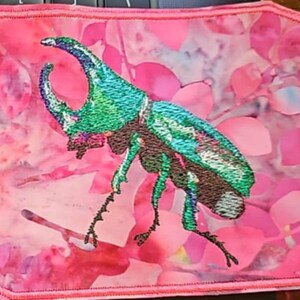 May include: Embroidered patch featuring a detailed beetle with iridescent green and blue accents. The insect is set against a pink floral background with a stitched border. This patch is suitable for jackets, bags, or other fabric items.