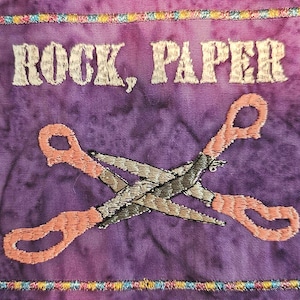 May include: Embroidered purple fabric with the words "ROCK, PAPER" in white thread. The image features a pair of crossed scissors with peach-colored handles and silver blades. The border is a colorful, stitched design.