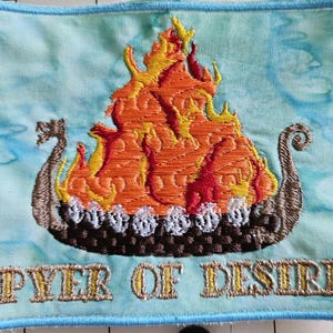 May include: Embroidered patch featuring a Viking ship engulfed in flames, with orange, yellow, and red hues. The ship is brown with white accents. The text "PYER OF DESIRE" is embroidered in gold lettering on a light blue background.