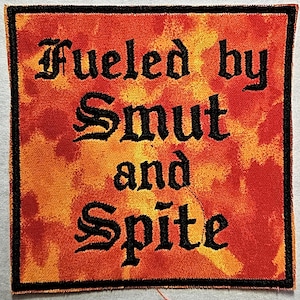 May include: Square patch with the text "Fueled by Smut and Spite" in black gothic lettering. The background is a blend of orange and red hues, with a black border.