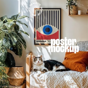 May include: A framed Bauhaus poster with a geometric eye design hangs above a bed. The poster features a blue eye, red and white accents, and the word "Bauhaus". A cat rests on the bed, with an orange pillow and decorative plants.
