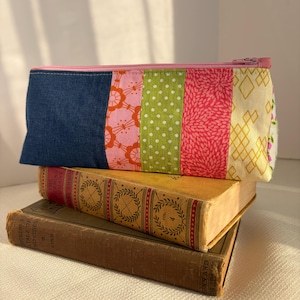 May include: A rectangular zippered pouch featuring a patchwork design of vertical fabric strips. The pattern includes a solid dark blue section, a pink floral print, a lime green polka dot section, a pink textured print, and a cream diamond pattern.