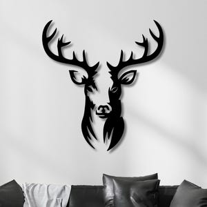 Puede incluir: Una pieza de arte mural de metal negro con forma de cabeza y cornamenta de ciervo. El diseño presenta una silueta estilizada y simétrica con bordes limpios y definidos, que muestra al animal de frente con una gran cornamenta ramificada.