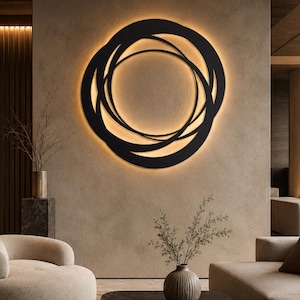 May include: A circular wall art piece featuring overlapping black rings with a warm, integrated backlight that creates a glowing halo effect against the wall. The design consists of multiple interconnected circular frames of varying thicknesses.