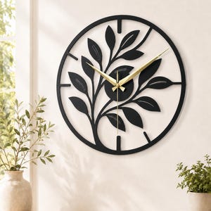 Puede incluir: Un reloj de pared de metal negro con un diseño de árbol. El reloj presenta un marco circular con una silueta de árbol recortada y manecillas doradas. El reloj está montado en una pared blanca, con elementos decorativos en primer plano.