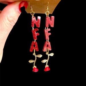May include: Red glittery dangle earrings spelling out the letters NFA, with gold-colored hooks and rose accents. The earrings are hanging from a brown card. The letters are in a bold, block style.
