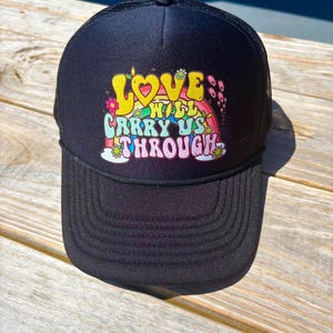 Könnte beinhalten: Schwarze Trucker-Cap mit gebogenem Schirm und Mesh-Rückseite. Auf der Vorderseite steht der Text "LOVE WILL CARRY US THROUGH" in farbenfrohen Retro-Lettern mit Blumen- und Regenbogenmotiven.