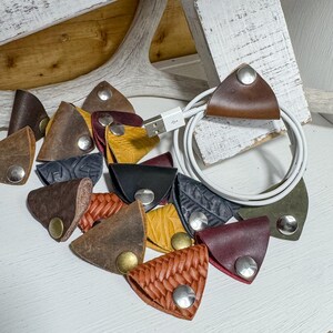 May include: A collection of leather cable organizers in various colors, including brown, black, yellow, and burgundy. Each triangular organizer has a metal snap closure. A white USB cable is looped through one of the organizers.
