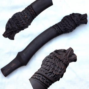May include: Dark brown, carved wooden handle with wolf head and geometric designs. The handle is curved and appears to be part of a larger tool or weapon. The wolf heads are intricately carved with open mouths.