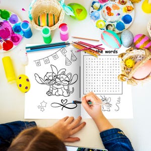 May include: A coloring book with a black and white illustration of two cartoon characters hugging. The book is open, and a hand is holding a pen. Easter eggs, paint, and art supplies surround the book.