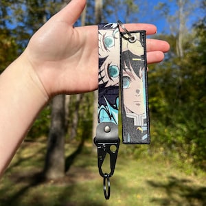 May include: A black lanyard with a cartoon character design. The lanyard features a black clasp and a split ring. The design includes a character with blue eyes and black hair. The background is a blurred outdoor scene.