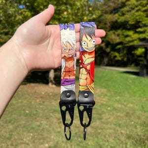 May include: Two colorful lanyards featuring anime characters. One lanyard has a white background with a character in white hair and purple pants. The other lanyard has a red background with a character in a red shirt and yellow pants. Both have black hardware.