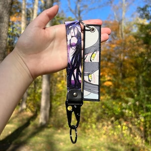 May include: A black lanyard with a purple and black anime character design and a matching keychain. The lanyard features a black clasp and a key ring. The keychain has a design of anime eyes.