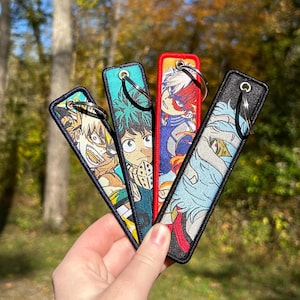 May include: Four embroidered keychains featuring anime characters. Each keychain has a different character design with vibrant colors and a metal keyring. The keychains are rectangular with a black border and are held in a hand.