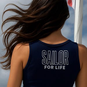 May include: Navy blue tank top with the words "SAILOR FOR LIFE" in white block letters. The top is ribbed and the model's hair is blowing in the wind. A flag is visible in the background.