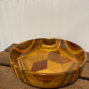 May include: A wooden salad bowl with a geometric design. The bowl features a mix of light and dark wood tones, with a starburst pattern in the center. The bowl is round with a slightly raised rim.