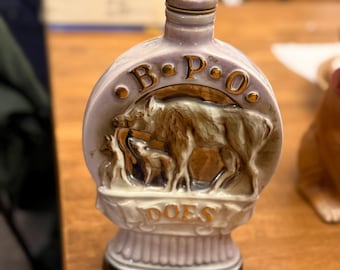 Op de afbeelding: Een keramische fles met een lichtpaarse glazuur. De fles heeft een reliëf van een wild zwijn en biggetjes, met de letters "B.P.O. DOES" in goud. De bovenkant heeft de woorden "PATRIOTISM, CHARITY, LOYALTY, LOVE".