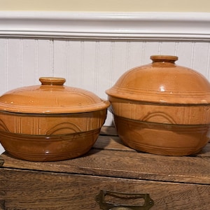 Peut inclure: Deux bols en céramique marron avec couvercles. Les bols ont une forme arrondie avec des motifs gravés décoratifs et une finition brillante. Les couvercles ont une petite poignée en forme de bouton. Les bols sont posés sur une surface en bois.