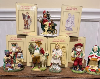 Op de afbeelding: Een verzameling kerstbeeldjes uit "The International Santa Claus Collection". De beeldjes beelden de kerstman, engelen en andere feestelijke figuren uit. De beeldjes zitten in hun originele dozen, met diverse ontwerpen en kleuren.