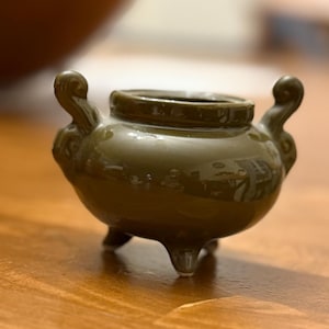 May include: A small, olive-green ceramic incense burner with three short legs and two curved handles. The burner has a glossy finish and a round opening at the top. The design is simple and elegant, suitable for home decor.