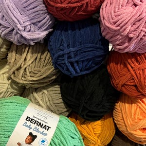 May include: A collection of colorful Bernat Baby Blanket yarn skeins in various shades, including green, purple, orange, and blue. The yarn is soft and plush, ideal for baby blankets and other craft projects. The label on the green skein reads "Bernat Baby Blanket".
